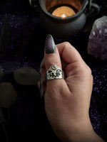 A hand with long dark gray manicured nails wearing a silver ring with a detailed witch silhouette and crescent moon design, set against a dark, mystical background with a cauldron and purple glitter.