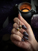 A hand with long dark metallic nails holds two small cone-shaped pendant earrings with gold and silver details against a dark, mystical background with a cauldron and purple crystal.