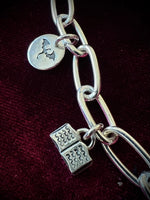 Silver chain bracelet with two charms, a dragon silhouette and a book, on a dark fabric background.