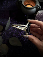 A hand with dark gray manicured nails holds a silver-toned ring featuring a large black onyx stone with a central gold inlay, set against a dark, mystical background with a cauldron, purple crystal, and leaves.