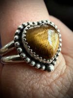 A close-up of a silver ring with a heart-shaped brown tiger's eye stone, worn on a finger.