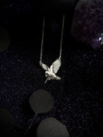 A silver-toned bird pendant necklace with spread wings, hanging against a dark glittery background with decorative leaves and a purple crystal.