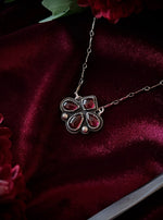 Silver necklace with a red gemstone pendant on a burgundy fabric background with flowers and a sword.