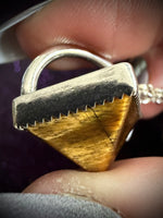 A close-up of a triangular pendant featuring a textured gold tiger's eye face and a dark silver top band, held by fingers.