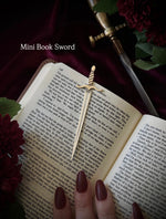 Open book with a sword-shaped bookmark and a hand holding it, on a dark background surrounded by flowers and a sword.