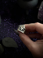 A hand with long dark nails holds a silver ring featuring a detailed moon and stars design, set against a dark background with purple glitter and crystals.