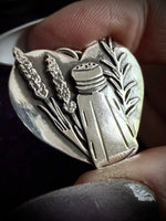 A close-up of a heart-shaped silver pendant held between fingers. The pendant features an engraved design of a salt and pepper shaker flanked by wheat stalks and herbs.