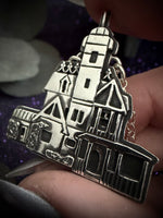 A close-up of a silver-toned house-shaped pendant being held by fingers against a dark, sparkly purple background.