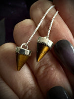 Two silver cone pendants with gold tiger's eye inlays on a silver chain, held in a person's hand with dark painted fingernails.
