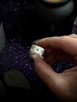 A hand holding a silver ring decorated with small floral or snowflake-like cutout patterns, set against a dark background with purple glitter.