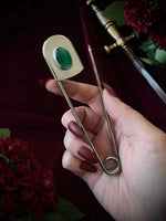 Giant stainless steel safety pin with a green gemstone on a dark burgundy fabric background with flowers and a sword.