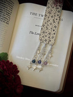 Three silver celestial bookmarks with star charms on an open book against a dark background.
