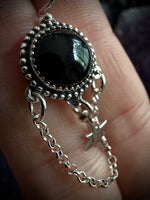 A close-up of a hand holding a silver-toned pendant necklace with a large round black stone and a dangling chain ending in a small star charm.