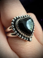 A close-up of a silver ring with a large black heart-shaped stone, worn on a finger.