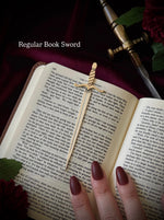 Open book with a sword-shaped bookmark and a hand holding it, on a dark background surrounded by flowers and a sword.