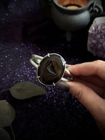 A hand holding a silver-toned cuff ring with a large oval dark agate stone, set against a dark purple glittery background with decorative leaves and crystals.