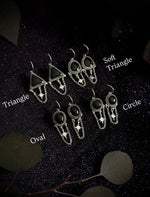 Four pairs of dangling earrings displayed on a dark textured background with leaf props. The earrings are labeled with text overlays reading Triangle, Soft Triangle, Oval, and Circle, showing different geometric and oval shapes with silver-tone chains and black stones.