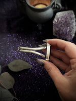 A hand with dark gray manicured nails holds a silver-toned metal ring with a unique multi-pronged design, set against a dark purple glittery background with decorative leaves, a cauldron, and an amethyst crystal.