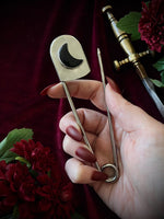 Hand holding a giant steel safety pin with a black crescent moon gemstone against a dark background with flowers and a sword.
