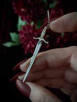 Silver sword-shaped earring held by a hand with a blurred floral background.