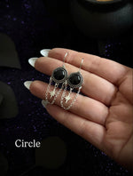 A pair of silver-toned dangle earrings with round black stones and dangling star charms, held on a person's hand against a dark glittery background. The word 'Circle' appears in the lower left.