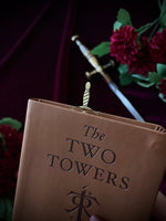 Leather book titled 'The Two Towers' with a sword bookmark sticking out of the top, against flowers and a sword in the background.