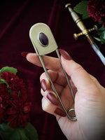 Hand holding a giant steel safety pin with an amethyst gemstone against a dark background with flowers and a sword.