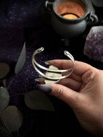 A hand with purple-gray nail polish holds a silver-toned metal cuff bracelet with dragon head details at both ends, set against a dark mystical background with a cauldron, amethyst cluster, and leaves.