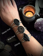Three silver wrap bracelets with different oval and oblong stone settings worn on a person's wrist, shown with text labels 'Skinny Oval', 'Wide Oval', and 'Oblong' against a dark purple glittery background with a candle and crystals.