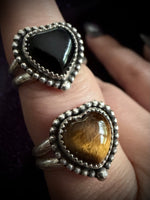 Two heart-shaped silver rings with textured bezels, one featuring a smooth black stone and the other a golden-brown tiger's eye stone, shown worn on a hand.