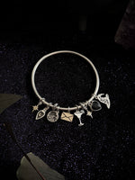 A silver charm bracelet with multiple small charms, including a heart, star, envelope, cocktail glass, and crescent moon, displayed on a dark glittery background.