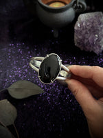 A hand holding a sterling silver cuff bracelet with a large, polished black onyx cabochon stone, set against a dark purple glittery background with decorative crystals and a cauldron.