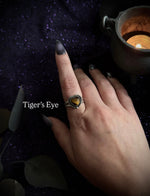A hand with dark gray nail polish wears a silver ring featuring a heart-shaped tiger's eye stone, set against a dark, mystical background with a burning candle and the text 'Tiger's Eye'.