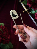 Hand holding a giant steel safety pin with a black crescent moon gemstone against a dark background with flowers and a sword.