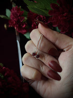 Hand holding a pair of dangly, silver earrings with red beads against a dark floral background.