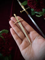 A hand holding a sword-shaped bookmark on a dark background surrounded by flowers and a sword.