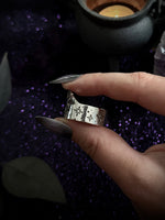 A hand with long dark manicured nails holds a wide silver band ring decorated with cutout snowflake and star patterns, set against a dark purple glittery background with a black cauldron prop.