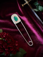 Giant stainless steel safety pin with a green gemstone on a dark burgundy fabric background with flowers and a sword.