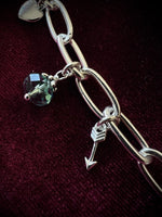 Silver chain bracelet with three charms, a heart, a gemstone, and an arrow, on a dark fabric background.
