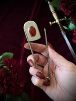 Hand holding a giant steel safety pin with a red gemstone against a dark background with flowers and a sword.