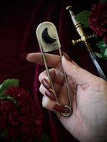 Hand holding a giant steel safety pin with a black crescent moon gemstone against a dark background with flowers and a sword.