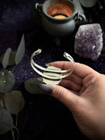 A hand with dark purple nail polish holds a silver-toned multi-band cuff bracelet against a dark, mystical background with a cauldron, amethyst cluster, and leaves.