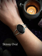 A person's wrist wearing a silver-toned skinny oval cuff bracelet with a black onyx stone, set against a dark background with a lit candle and purple crystals. The text 'Skinny Oval' is visible in the lower left corner.