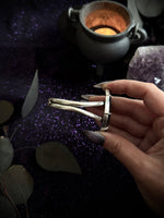A hand with long nude manicured nails holds a silver triple-halo ring against a dark, mystical background featuring a glowing cauldron, purple glitter, and crystals.