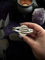 A hand holding a silver bracelet against a dark, mystical background with a cauldron, purple crystal, and leaves.