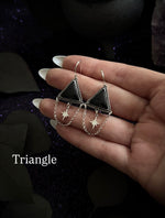 A pair of silver-toned triangle dangle earrings with black triangular stones and dangling star charms, held in a person's hand against a dark decorative background. The word 'Triangle' appears as text in the lower left.