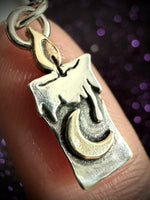 A close-up of a silver pendant shaped like a melting candle with a crescent moon, resting on a person's finger against a dark purple bokeh background.