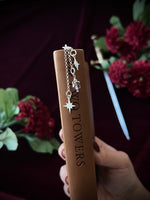 Hand holding a leather book with silver star charms hanging down on the binding, surrounded by flowers and a sword in the background.