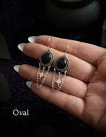 A pair of oval drop earrings with black oval stones and silver dangling chain charms, held in a person's hand against a dark glittery background. The word 'Oval' is printed in the lower-left corner.