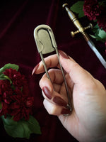 Hand holding the backside of a giant steel safety pin against a dark background with flowers and a sword.